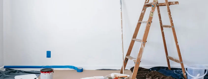 A room being prepped for painting. There's a bucket of paint, some painters tape on the wall, a paint roller leaning against the wall, and a ladder.