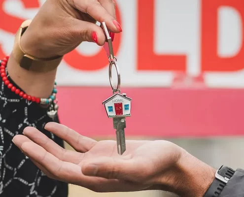 A real estate agent handing a house key to a homeowner in front of a "Sold" sign