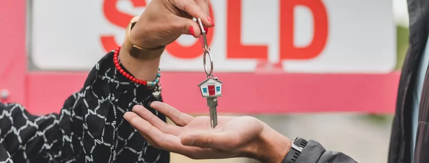 A real estate agent handing a house key to a homeowner in front of a "Sold" sign