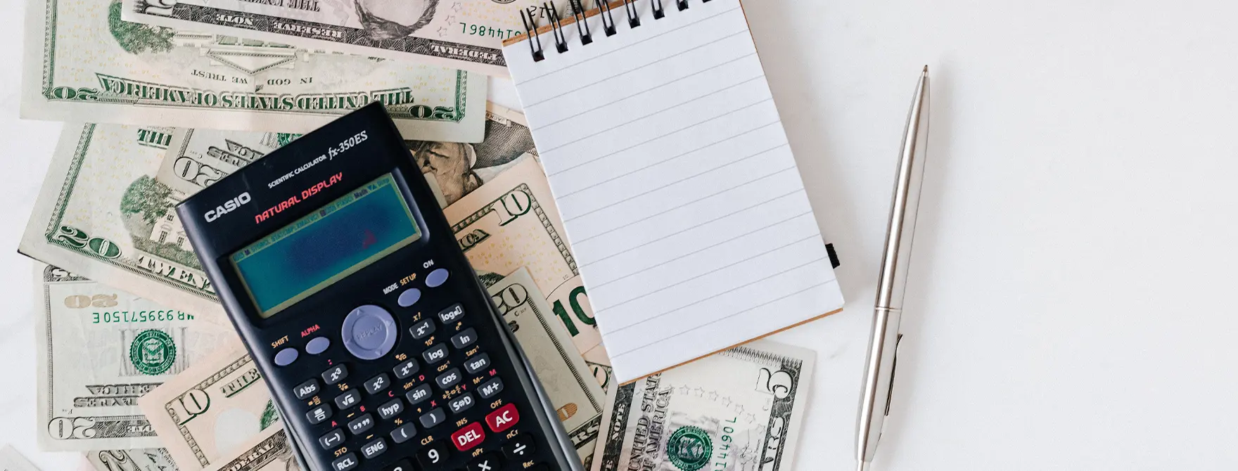 A calculator, notepad, and pen resting on a pile of cash