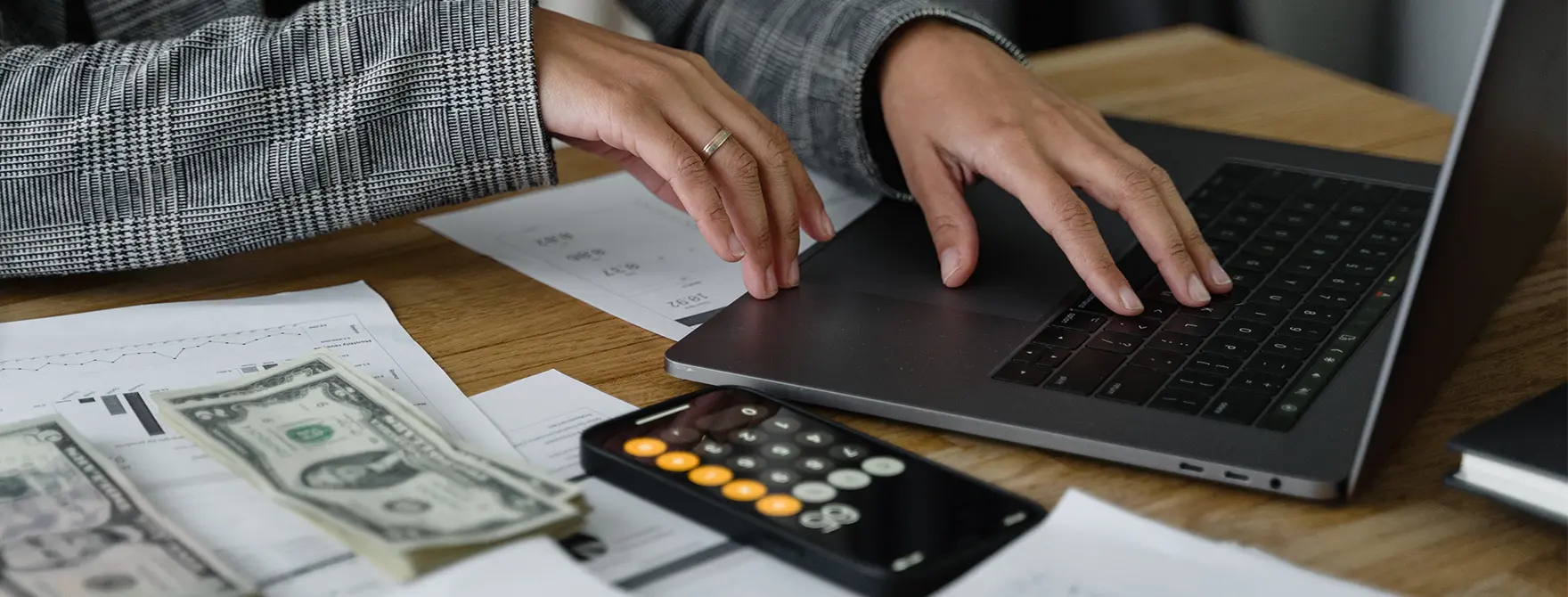 Someone typing on a laptop with a calculator, paperwork, and piles of money nearby