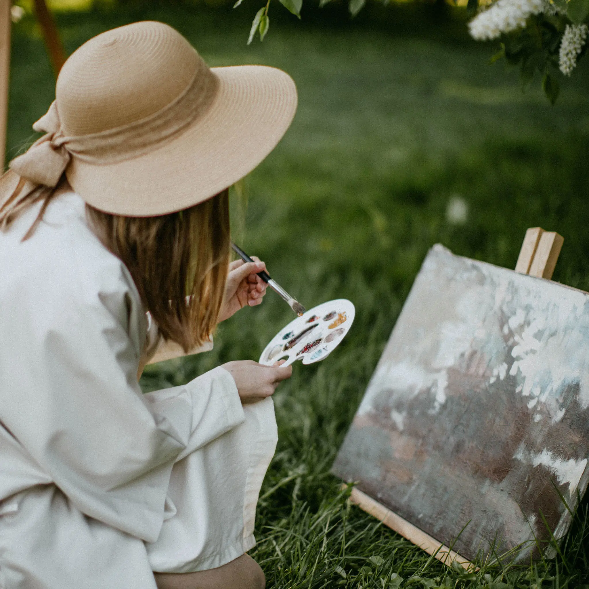 A woman wearing a large hat and painting on a canvas in the grass.