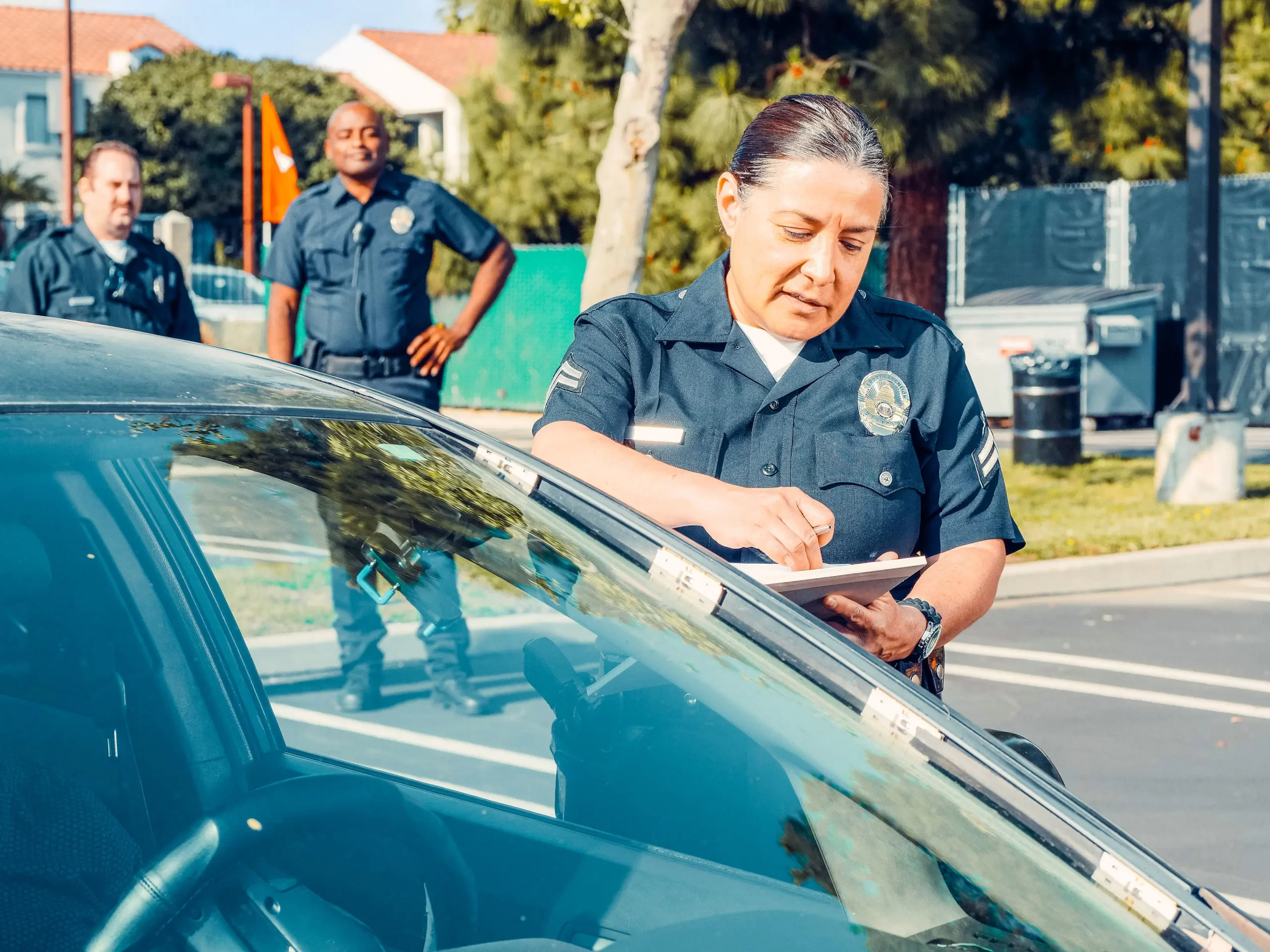 A female cop writing a ticket while two other cops wait in the background.