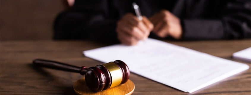 Close up on of a gavel resting on a sound block while a judge signs papers in the background.