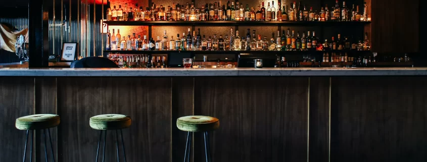 A bar with green barstools in front and several rows of alcohol in the back