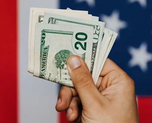 A hand holding American currency in front of an American flag