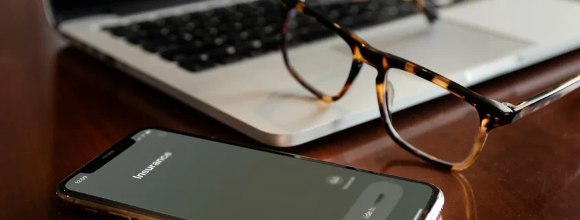 A cell phone with an incoming call from an insurance company, resting on a laptop with a pair of glasses