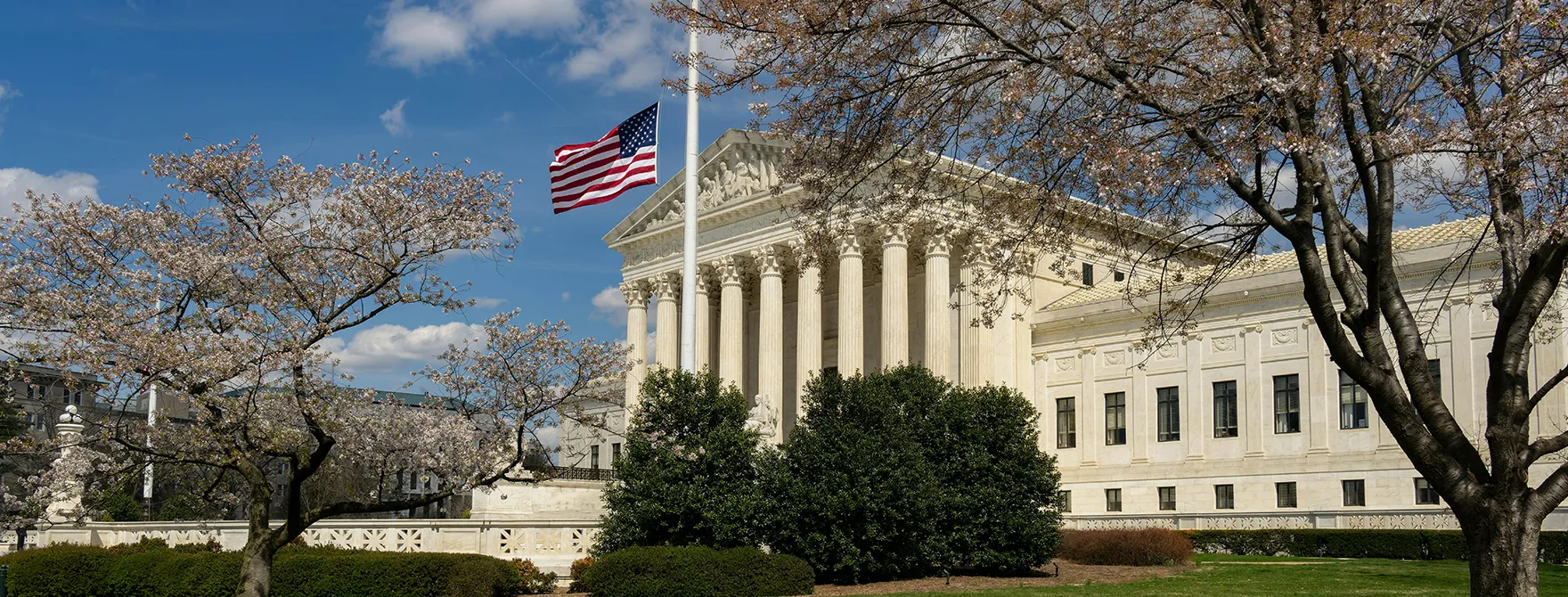 Supreme Court Header A picture of the courthouse where the Supreme Court of the United States meets framed by spring trees and an American flag.